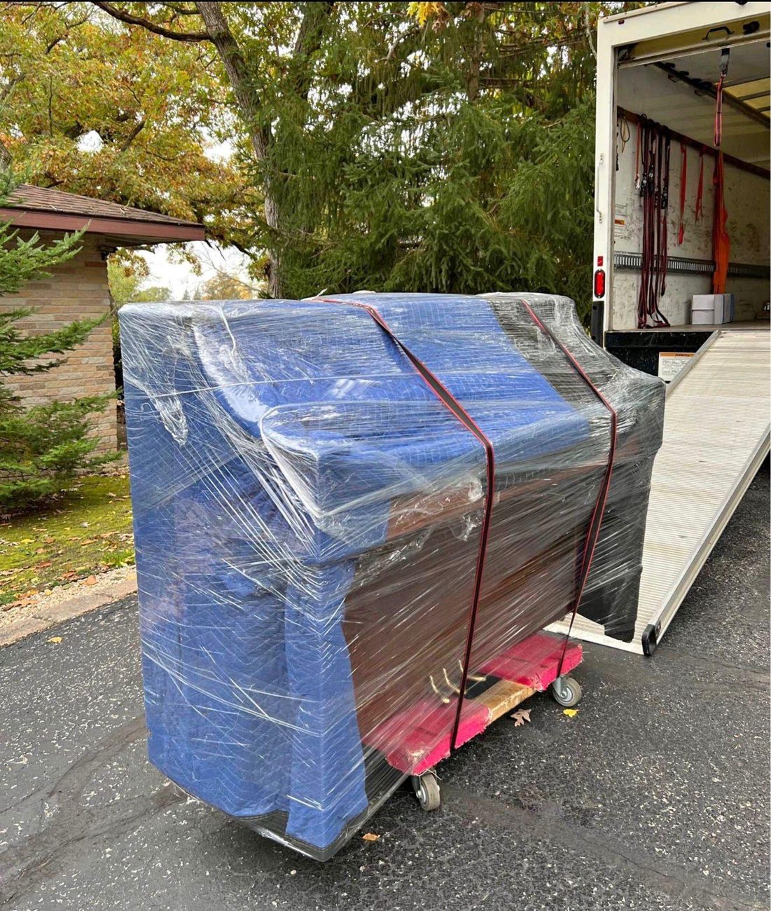 Protected upright piano wrapped in moving blankets and shrink wrap on a dolly beside a moving truck
